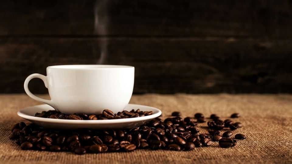 Steaming white coffee cup surrounded by roasted coffee beans on a rustic burlap surface.