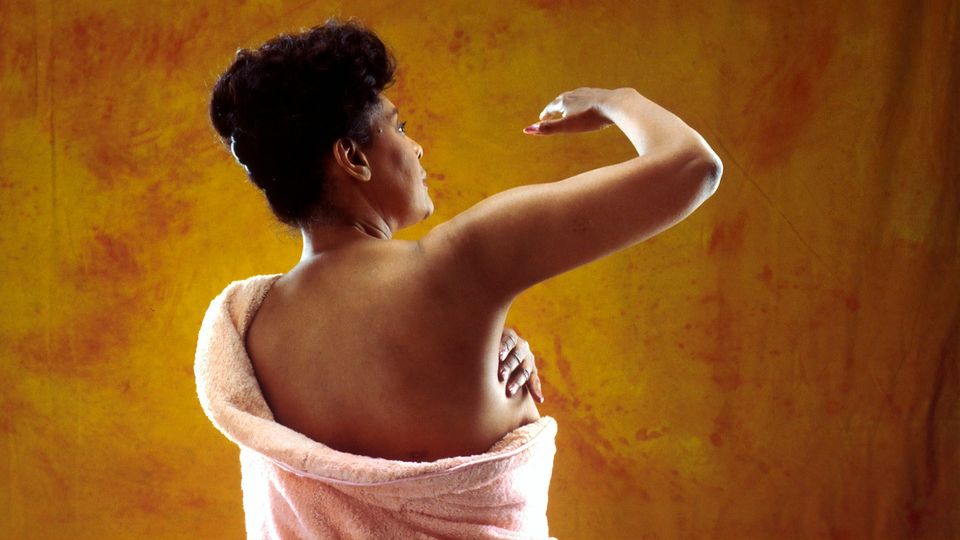 Woman wrapped in a towel performing a breast self-exam with her arm lifted, against a warm background.