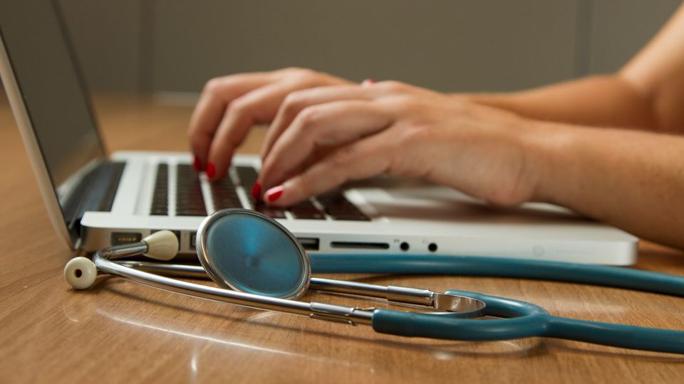 Healthcare professional typing on a laptop with a stethoscope on the desk.