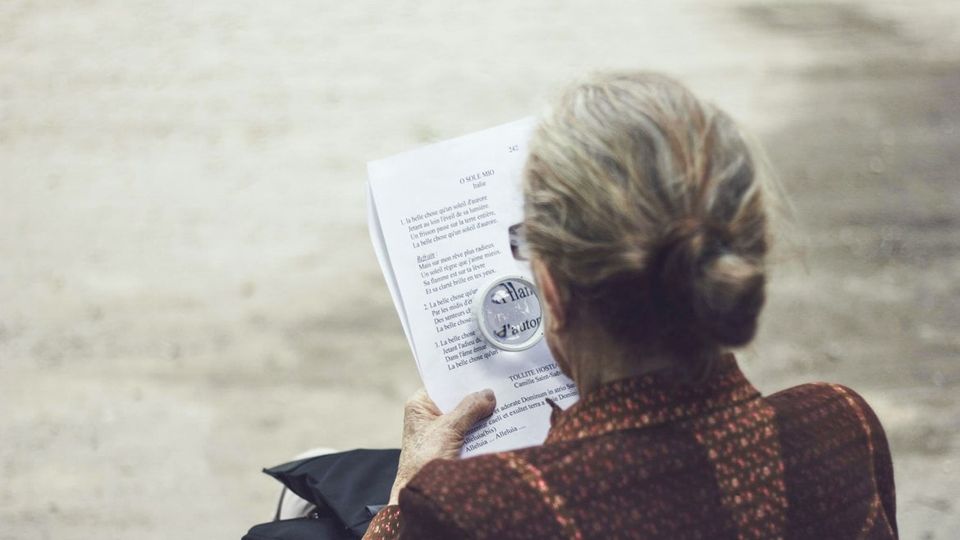 An elderly woman sits outside, reading a poem using a magnifying glass.