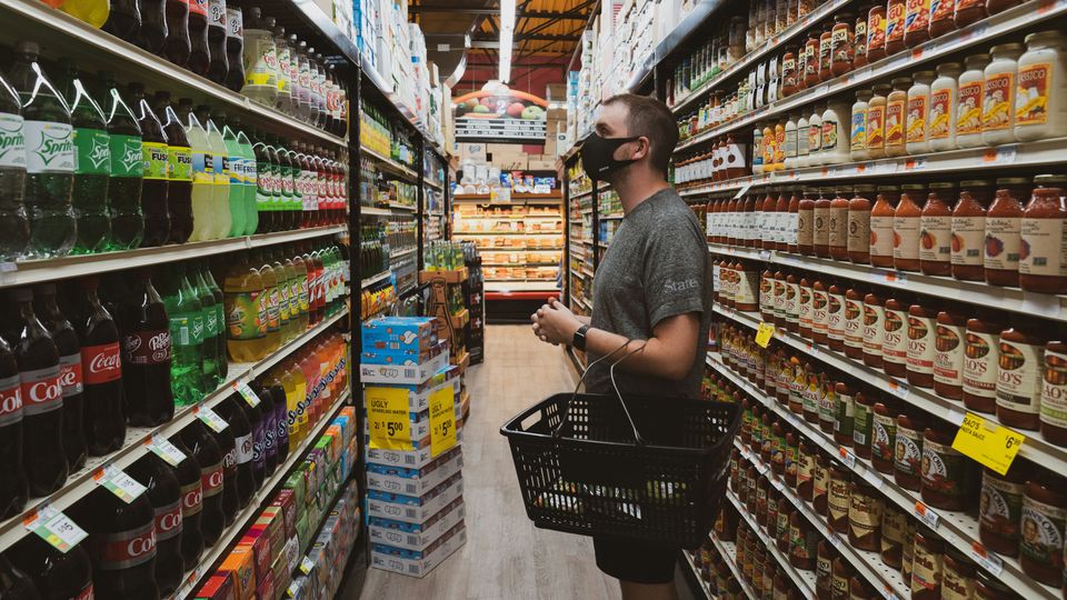 A man standing in an aisle at a grocery store, looking at shelves of soft drinks and sauce jars.