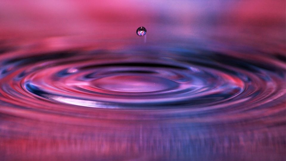 Colorful water droplet suspended, with pink and blue reflections on surface of liquid.