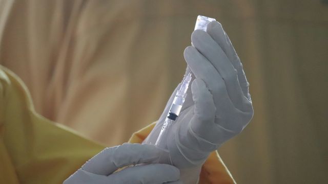 A gloved healthcare worker drawing clear liquid from a glass vial into a syringe. 