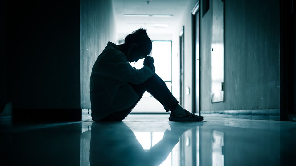 Woman sat against a wall in a dark corridor, with her knees up to her chest, holding her head in her hands.