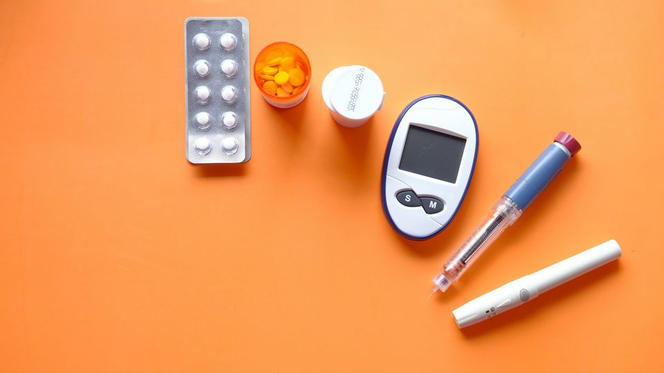 Birds-eye view of a pill sachet and container, a glucose meter, and diabetes medications on an orange background.