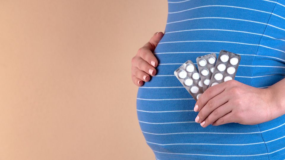 Pregnant woman holding medication blister packs against a neutral background.