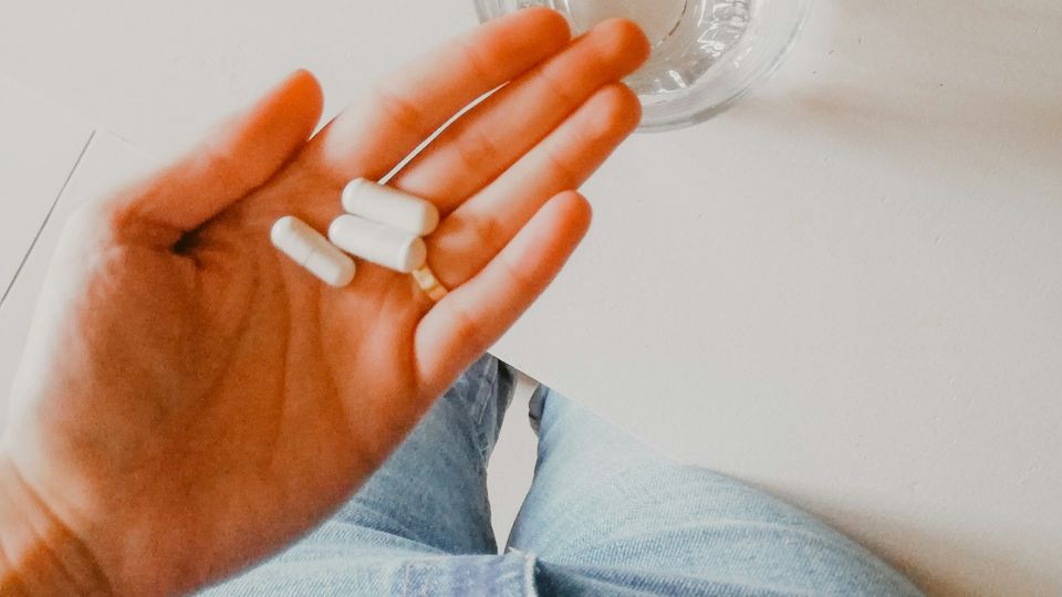 Hand holding several capsules and pills beside a glass of water, against a plain white background.