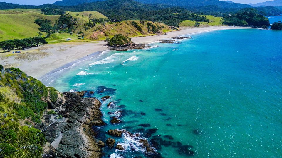 Aerial view of a turquoise beach with rocky shoreline, rolling hills, and coastal mountains in the background.