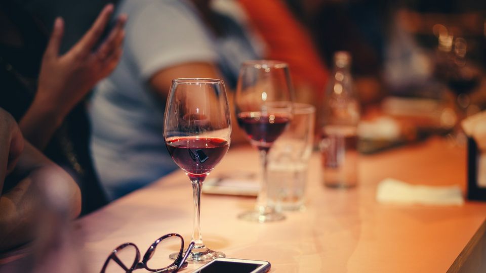 Glasses of red wine on a restaurant table with people socializing in the background.