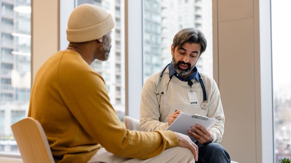 Clinician wearing a stethoscope and holding a clipboard, sat opposite a patient in a consulting room.