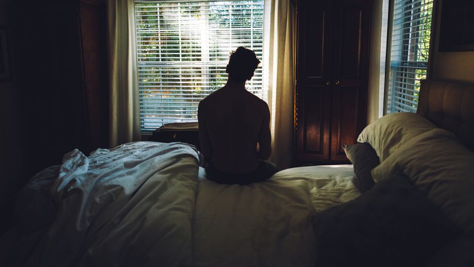 Person with depression sitting alone on a bed in a dark bedroom.