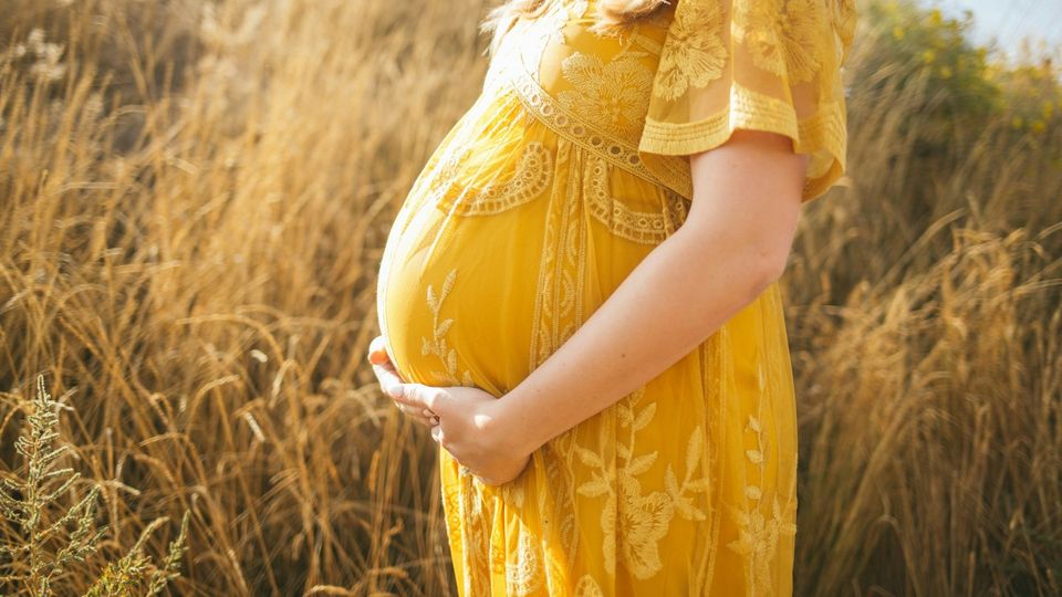Pregnant woman in a yellow dress holding her baby bump in a sunlit field.