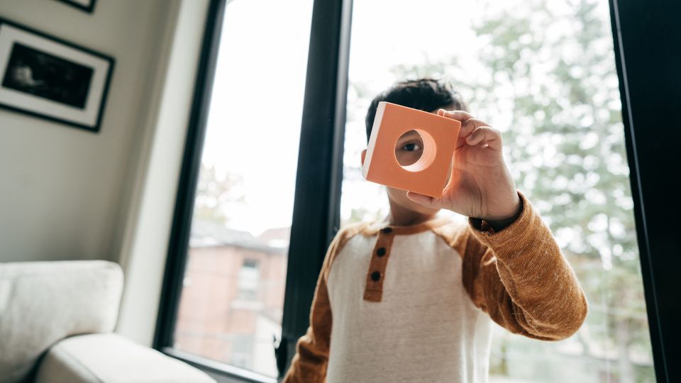 Child holds a square toy block up to their eye by a bright window.