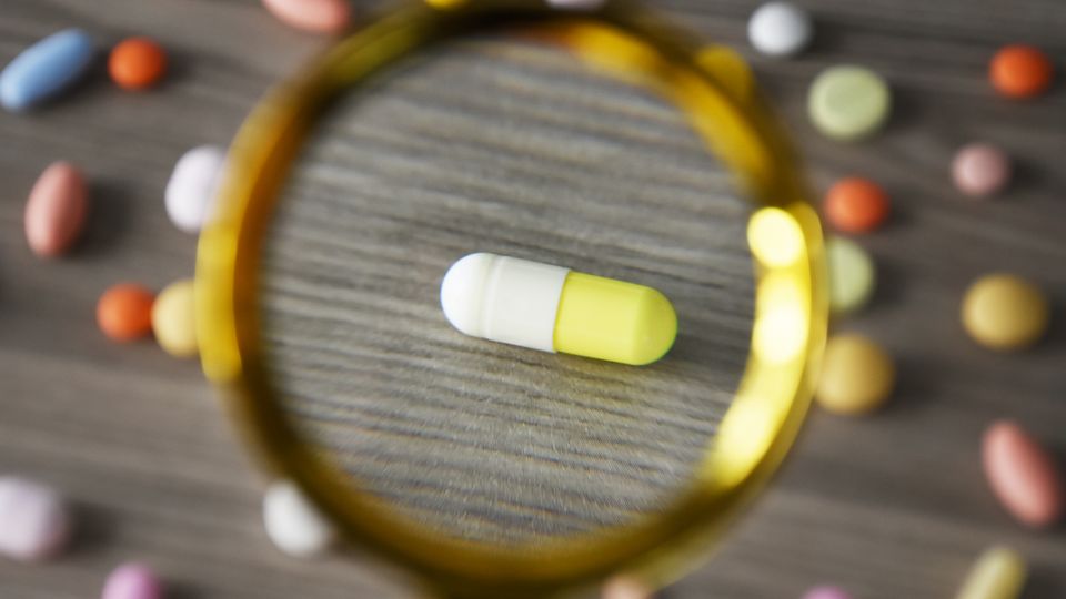 Capsule drug under a magnifying glass with colorful pills scattered nearby.