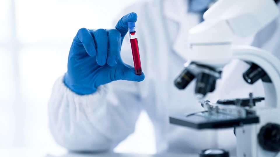 Scientist wearing a lab coat and gloves, holding a test tube full of blood in a laboratory setting.