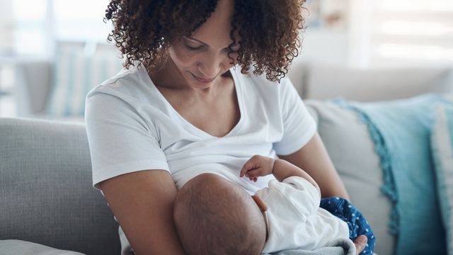 A woman breastfeeds a baby while sitting on a sofa. 
