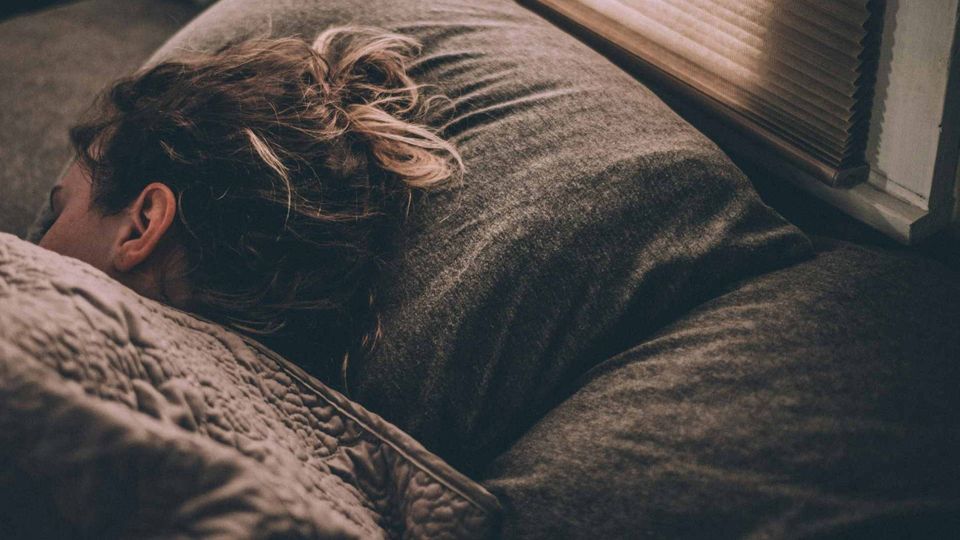A woman with messy hair sleeps on a dark pillow under a textured blanket, focusing on the act of sleep.