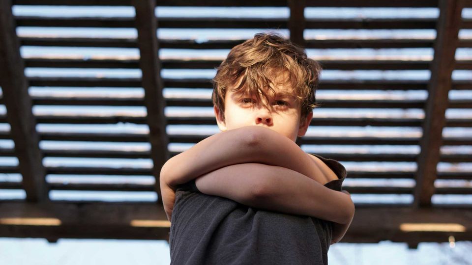 A young boy stands with his arms tightly crossed over his chest, looking directly forward.