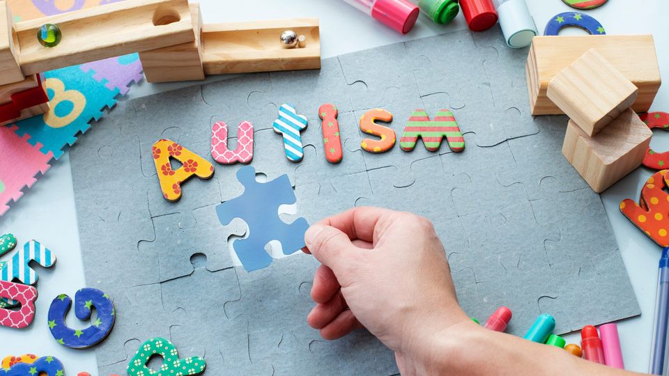 A hand placing the final piece into a gray jigsaw, with letters of various colors and patterns spelling "autism" above.