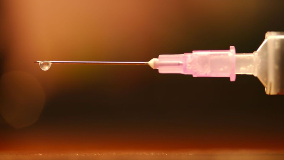 Close-up of a needle attached to a syringe with a droplet of fluid being released, against an orange background.