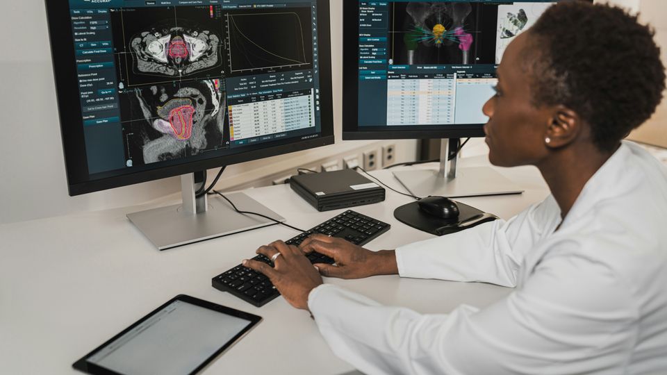 Researcher wearing a laboratory coat, sat at a desk analyzing medical scans on dual computer monitors.