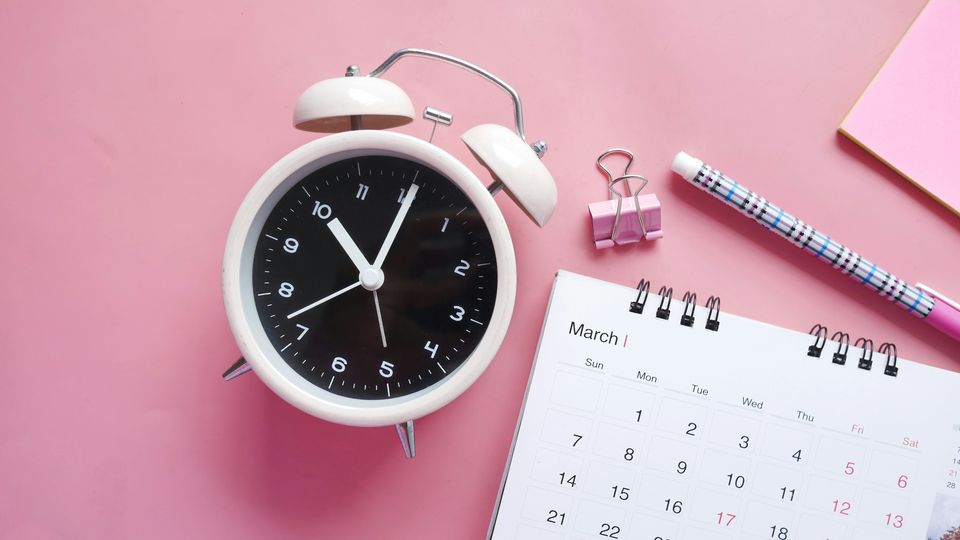Birds-eye view of an alarm clock, calendar, and stationary laid on a flat pink surface.