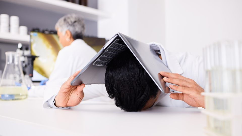 Researcher resting their head under a laptop in a laboratory, illustrating workplace stress.