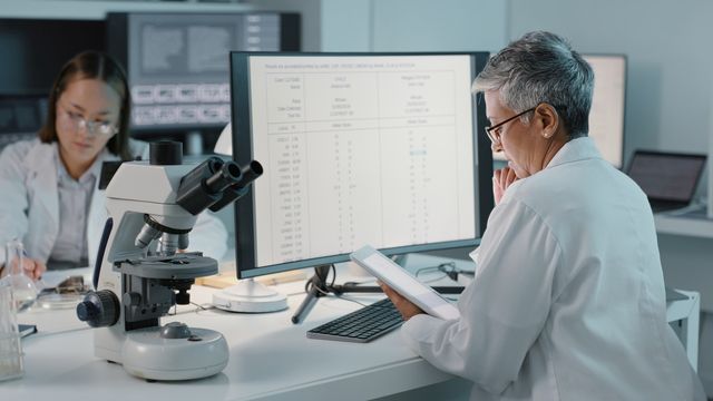 Pathologist sat at a desk looking at a tablet screen, with a large monitor and microscope in front of her. 