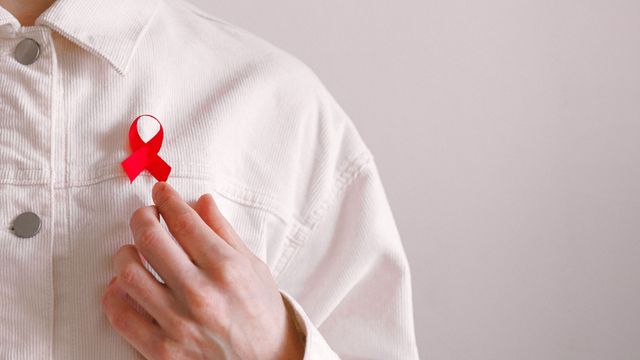Close-up of a red-ribbon pin on the front of a mans shirt. He is reaching one hand up to touch it. 
