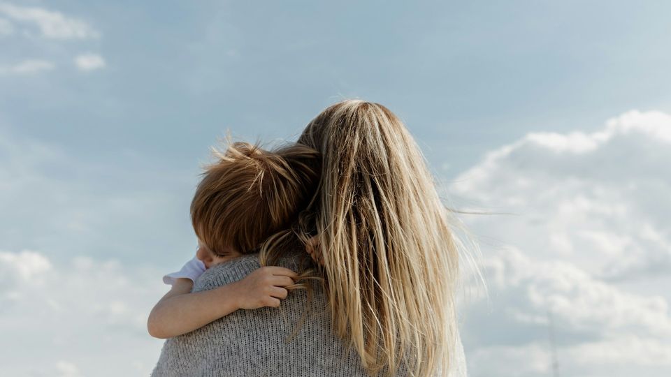 Woman holding and comforting a young child outdoors beneath a cloudy sky.