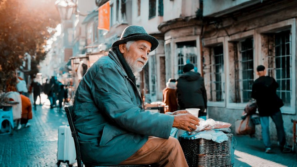Older man sitting on a city street holding a cup.