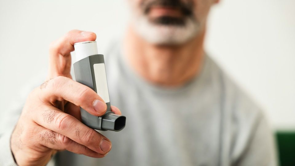 Man wearing a gray jumper holding an asthma inhaler up to the camera.