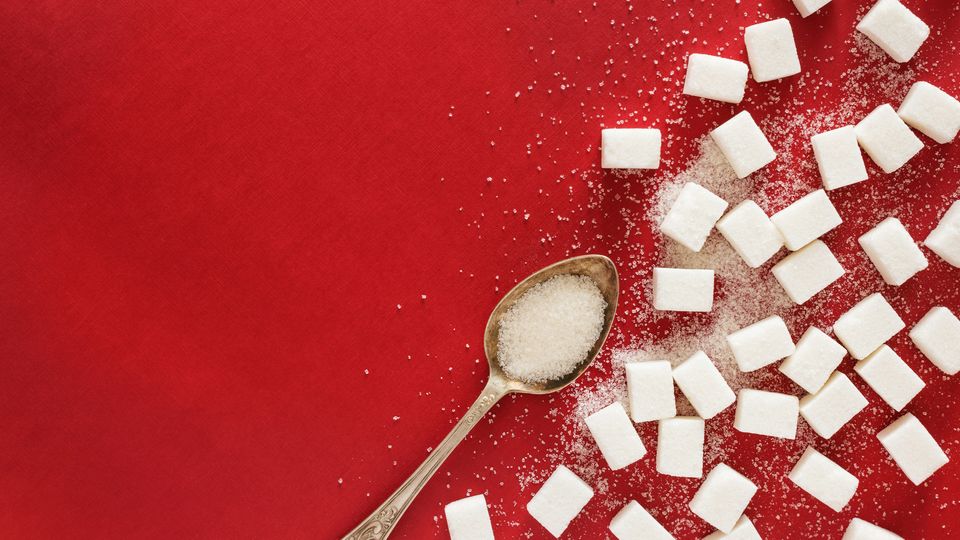 A teaspoon full of sugar laid next to sugar cubes on a red background.