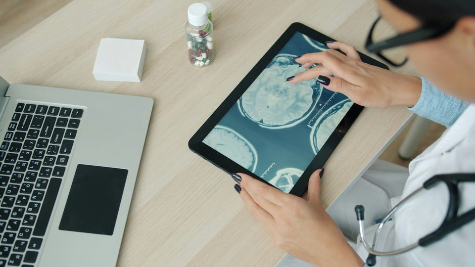 Doctor wearing a stethoscope reviewing brain scans on a tablet, with a bottle of pills and laptop next to her.