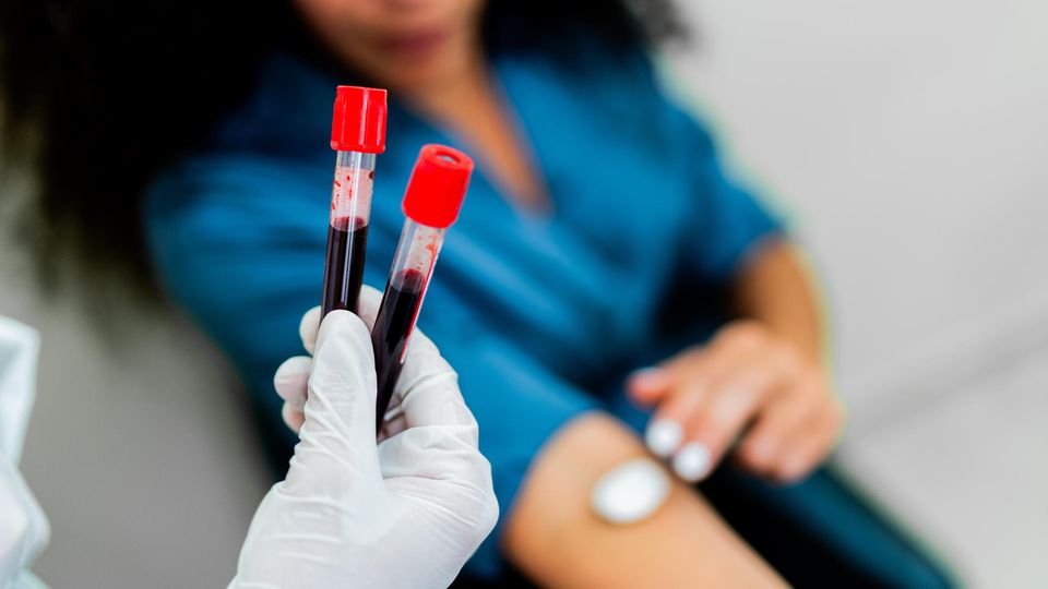 Gloved hand holding two test tubes containing blood samples. In the background, a woman is pressing a cotton ball to her inner elbow.