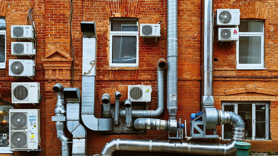 The side of a red brick building, covered in metal pipes and white air conditioning units.