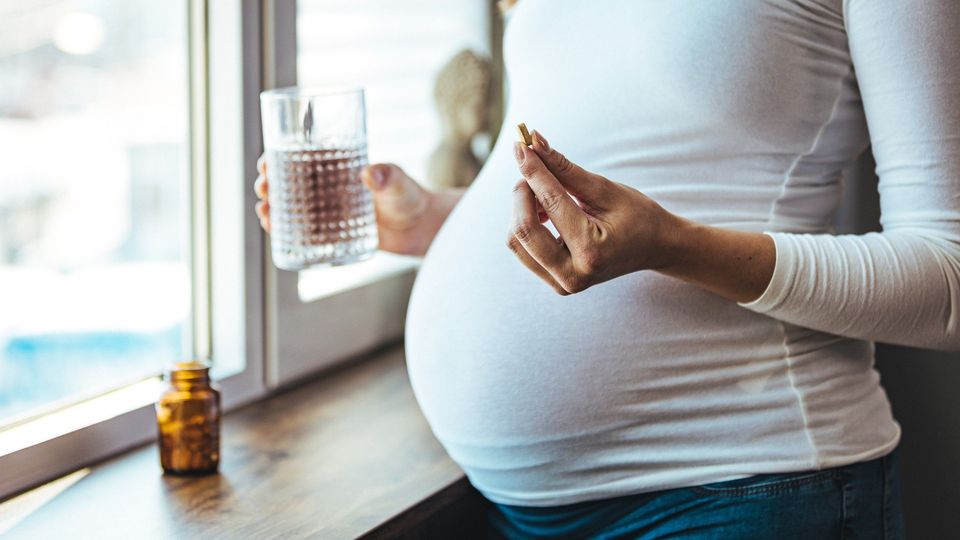 Mid-section of a pregnant person holding a capsule in one hand and a glass of water in the other hand.