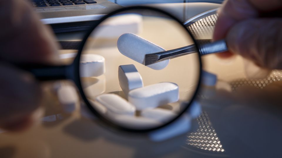 Magnified white tablets held by tweezers, representing drug inspection or pharmaceutical quality control.