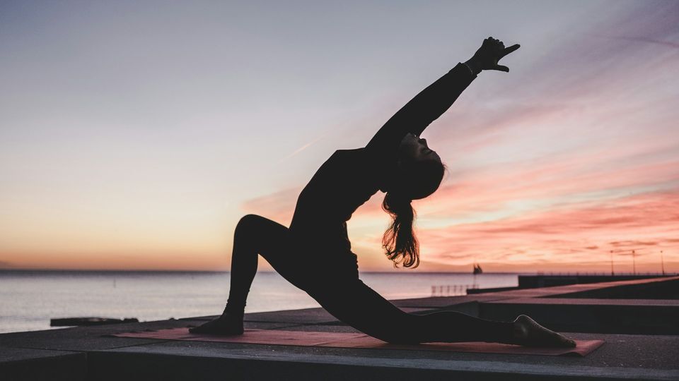 A woman doing yoga on a mat, arching her back with arms stretched upward, as the sun sets in the background.