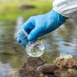 Gloved hand collecting a water sample for testing from a river in a glass round-bottom flask. 