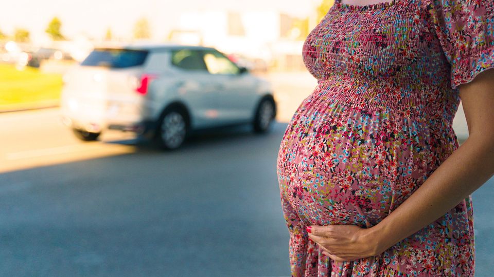 Person holding a visibly pregnant belly in a patterned dress, standing beside a road with cars in the background.