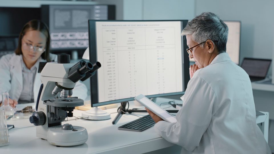 Scientist in a laboratory coat sat looking at a tablet, with a large monitor in front of her and a microscope next to her.