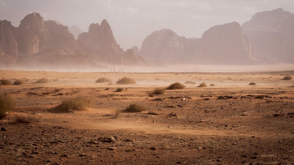 Dusty desert valley with scattered shrubs and rocky cliffs.