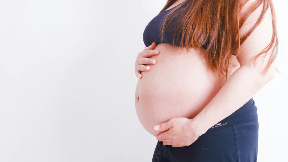 Mid-section of a pregnant woman holding her baby bump, stood next to a white background.