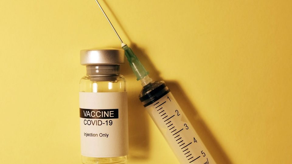 COVID-19 vaccine vial with a syringe next to it, placed on a yellow background.