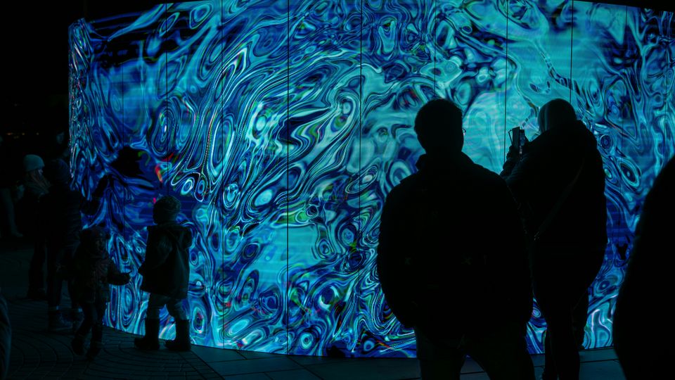 Silhouettes of people standing in front of a large LED screen in an art gallery.