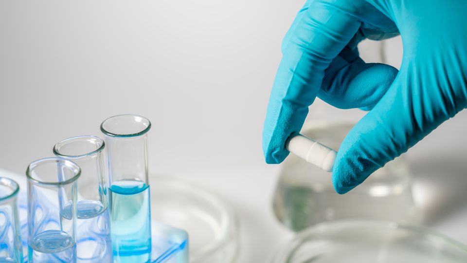 Gloved hand holding a capsule drug beside laboratory test tubes filled with blue liquid.