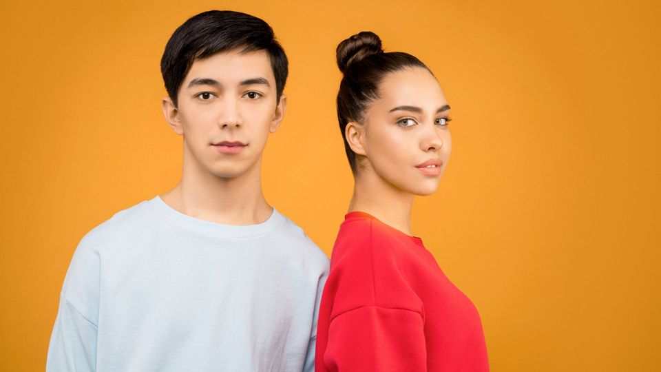 A man and woman standing together against an orange background, looking at the camera.