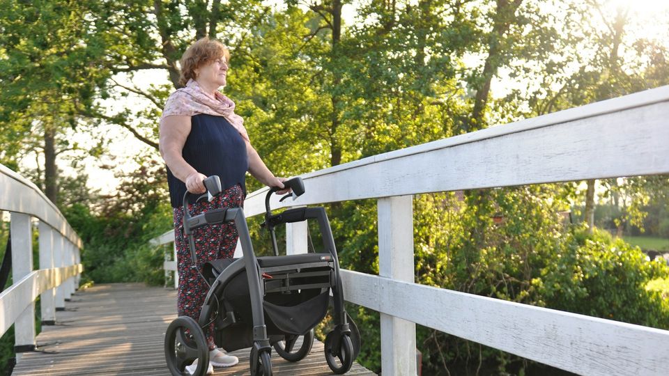Older woman using a rollator walker while standing on a wooden bridge in a park.