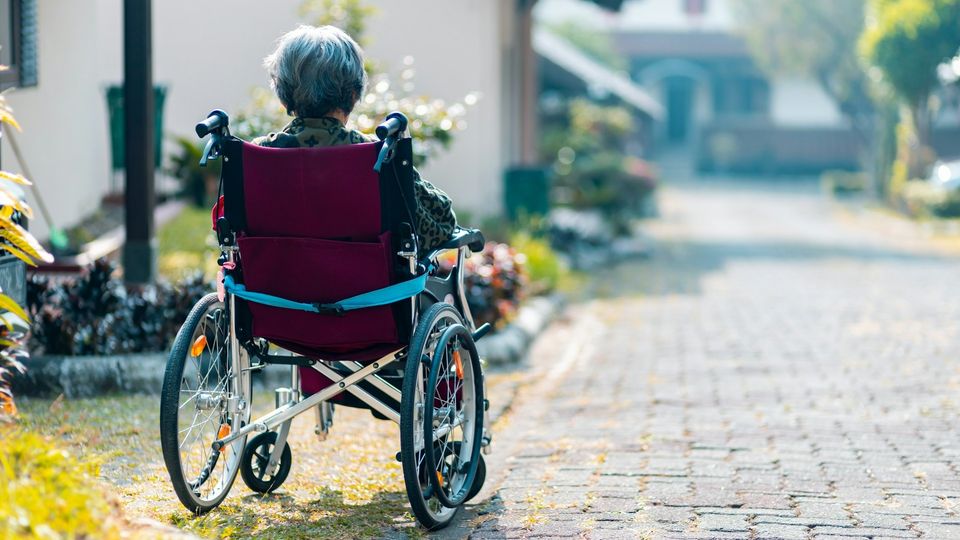 Older adult using a wheelchair on a sunlit residential pathway.